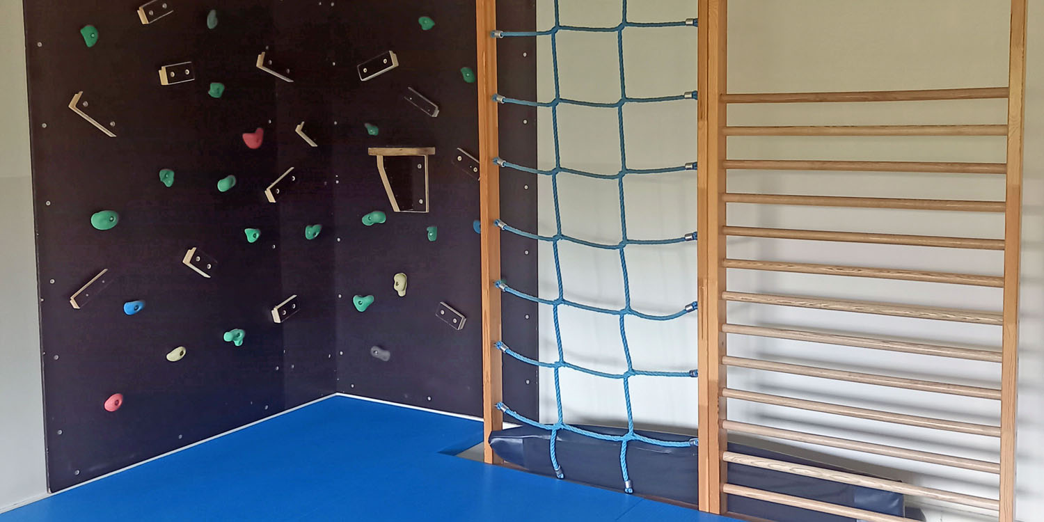 In einem Sportraum sind an der Wand eine Sprossenwand, eine Wand mit Seilen und eine Kletterwand. Im Vordergrund eine große Sportmatte.