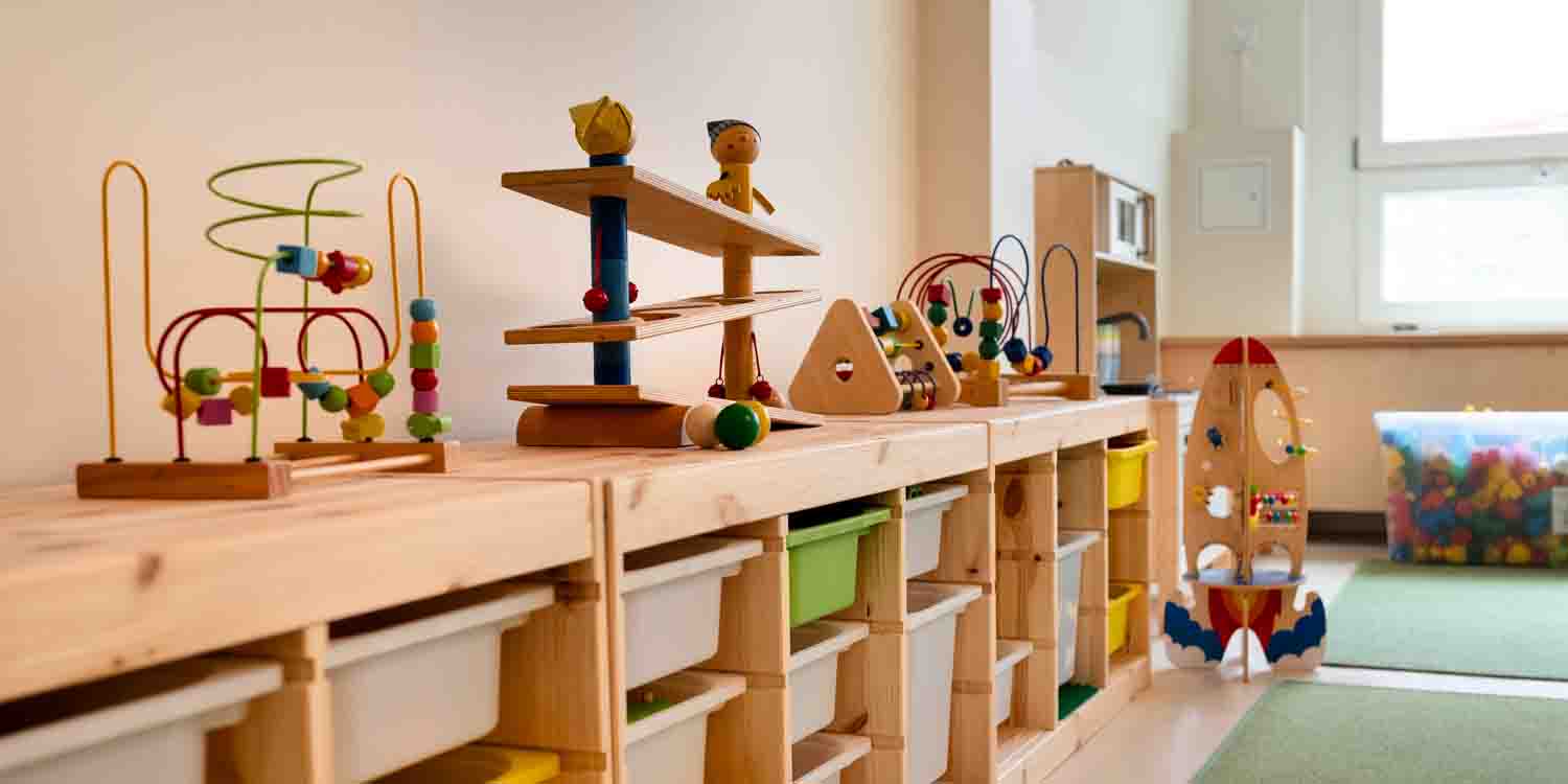 An der Wand eines Grupepnraumes sreht ein Regal mit Boxen. Auf dem Regal stehen Spielmaterialien aus Holz. Unter einem Fenster steht eine Kiste mit Legosteinen.