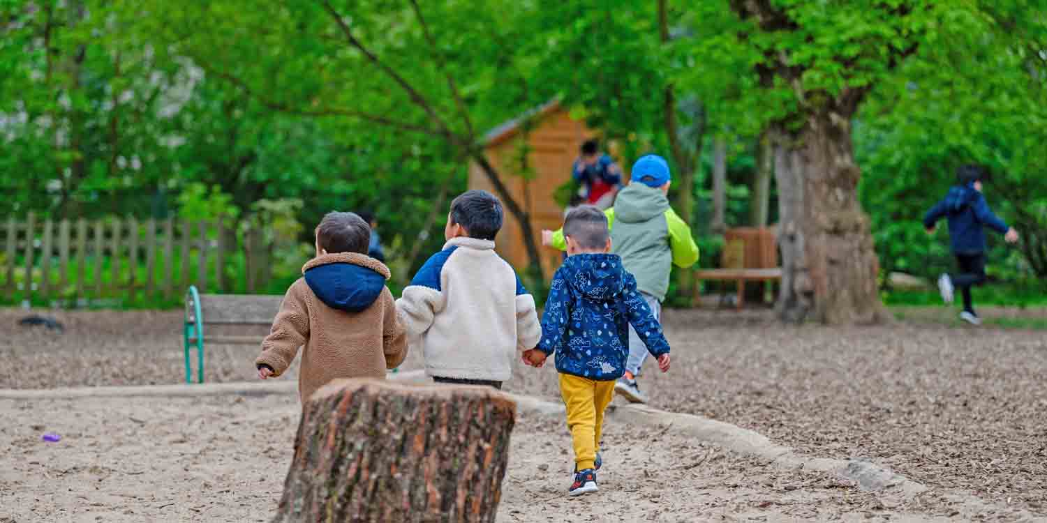 Kinder laufen in einem Kitagarten zu einer kleinen Holzhütte.