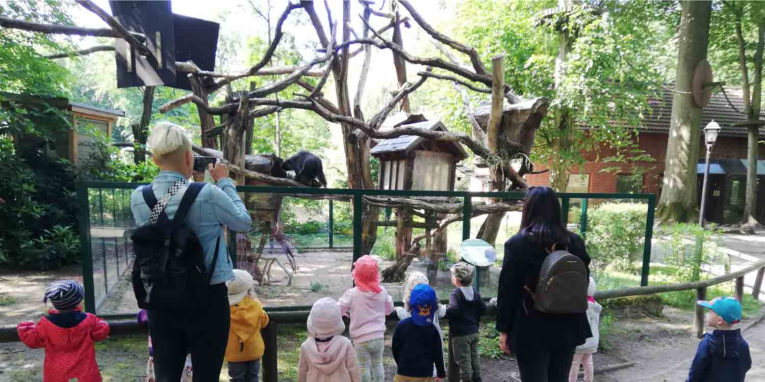 Kinder und zwei pädagogische Fachkräfte stehen vor einem Affengehege mit großen Klettermöglichkeiten.
