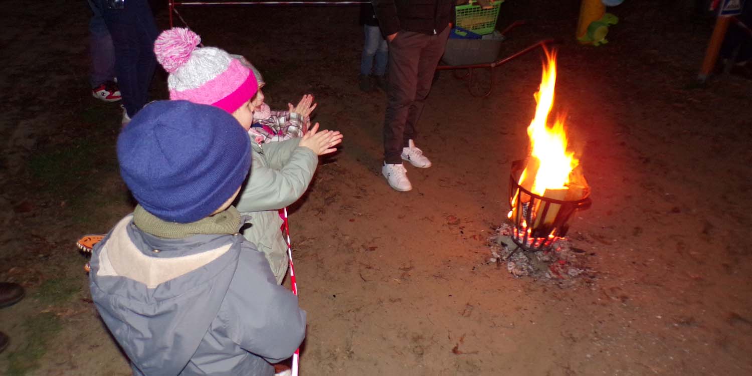 In einer Feuerschale in der Mitte eines Kreises brennt ein kleines Feuer. Kinder stehen hinter einem Absperrband und halten die Hände in Richtung Feuer.