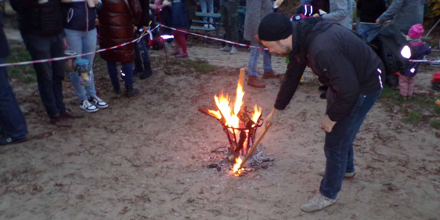 Ein Mann entfacht ein Feuer in einem Feuerkorb. Um ihn herum stehen Kinder und Eltern.