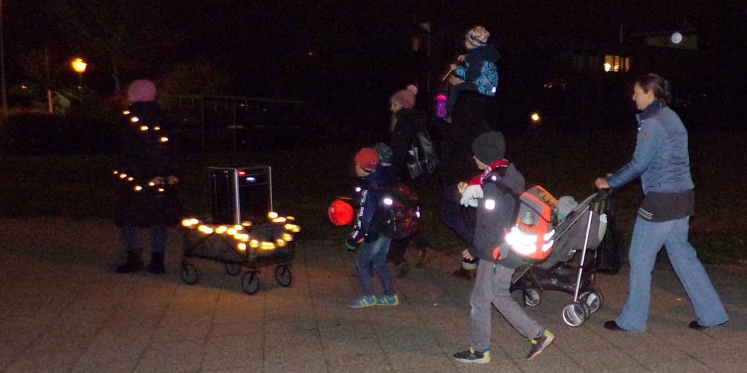 Mütter mit kleinen Kinden an der Hand und Kinderwagen laufen auf einer dunklen Straße, die mit Lampions beleuchtet wird.