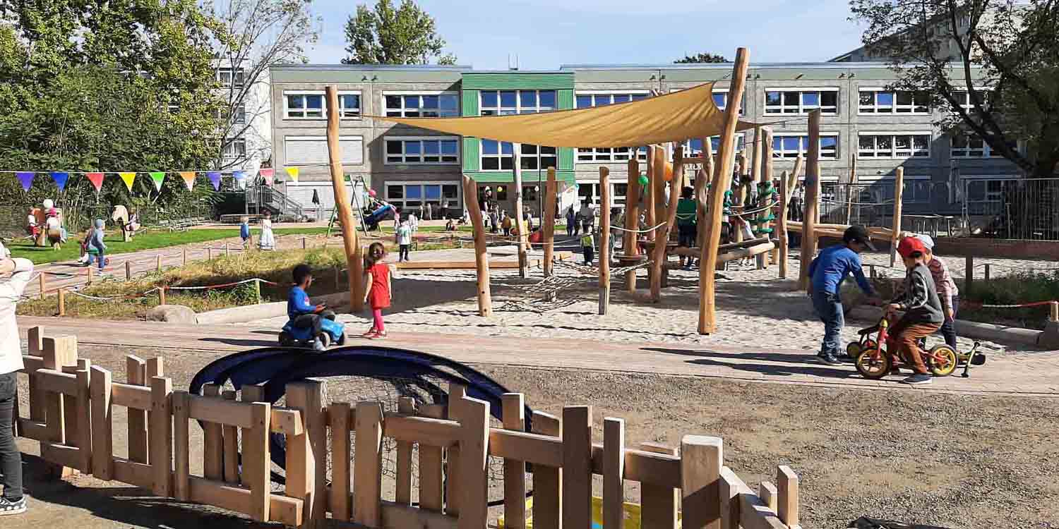 In einem großen Kita-Garten laufen Kinder und fahren auf Laufrädern auf den Wegen. Im Hintergrund steht ein dreigeschossiges Kitagebäude, davor der Spielplatz mit Holzklettergerüsten. Im Vordergrund ein kleiner Zaun aus Holz, der den Kleinkindbereich abgtrennt.