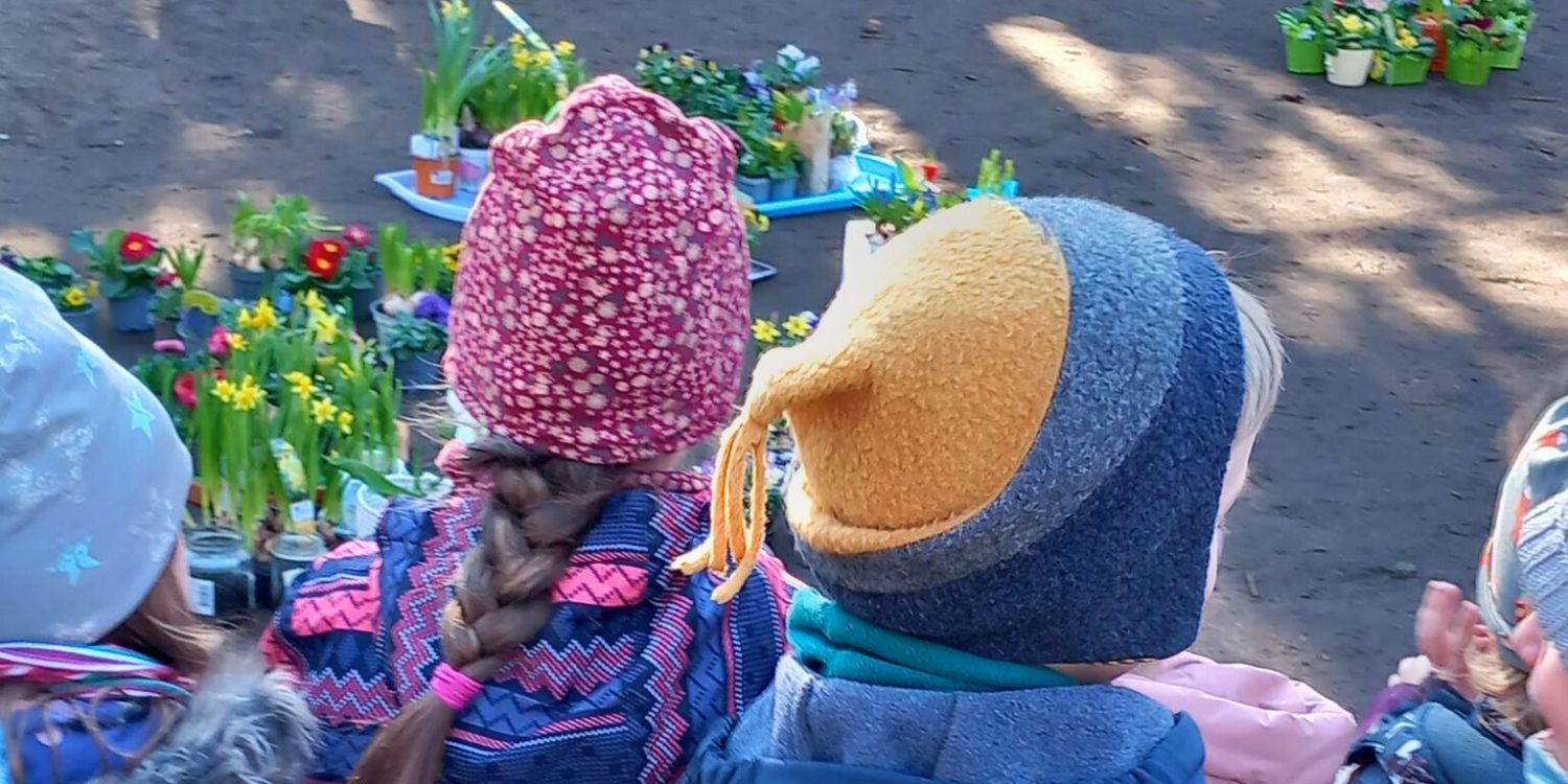 Zwei Kinder stehen inmitten von Blumentöpfen mit Frühlingsblühern.